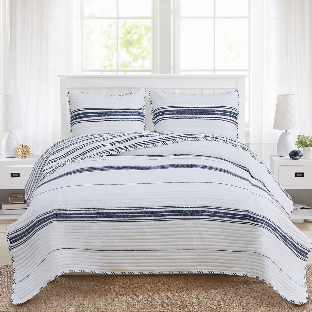 A reversible striped quilt set with a modern design, featuring navy and white stripes, displayed on a bed with two accompanying pillows. The quilt is draped over the bed with a visible white and navy striped pattern on both sides.