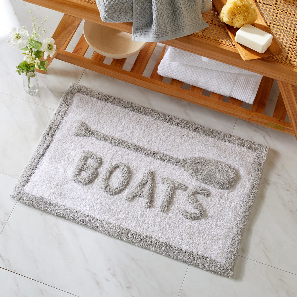 Gray doormat with 'BOATS' text on a bathroom floor, next to a wooden shelf with bath items.