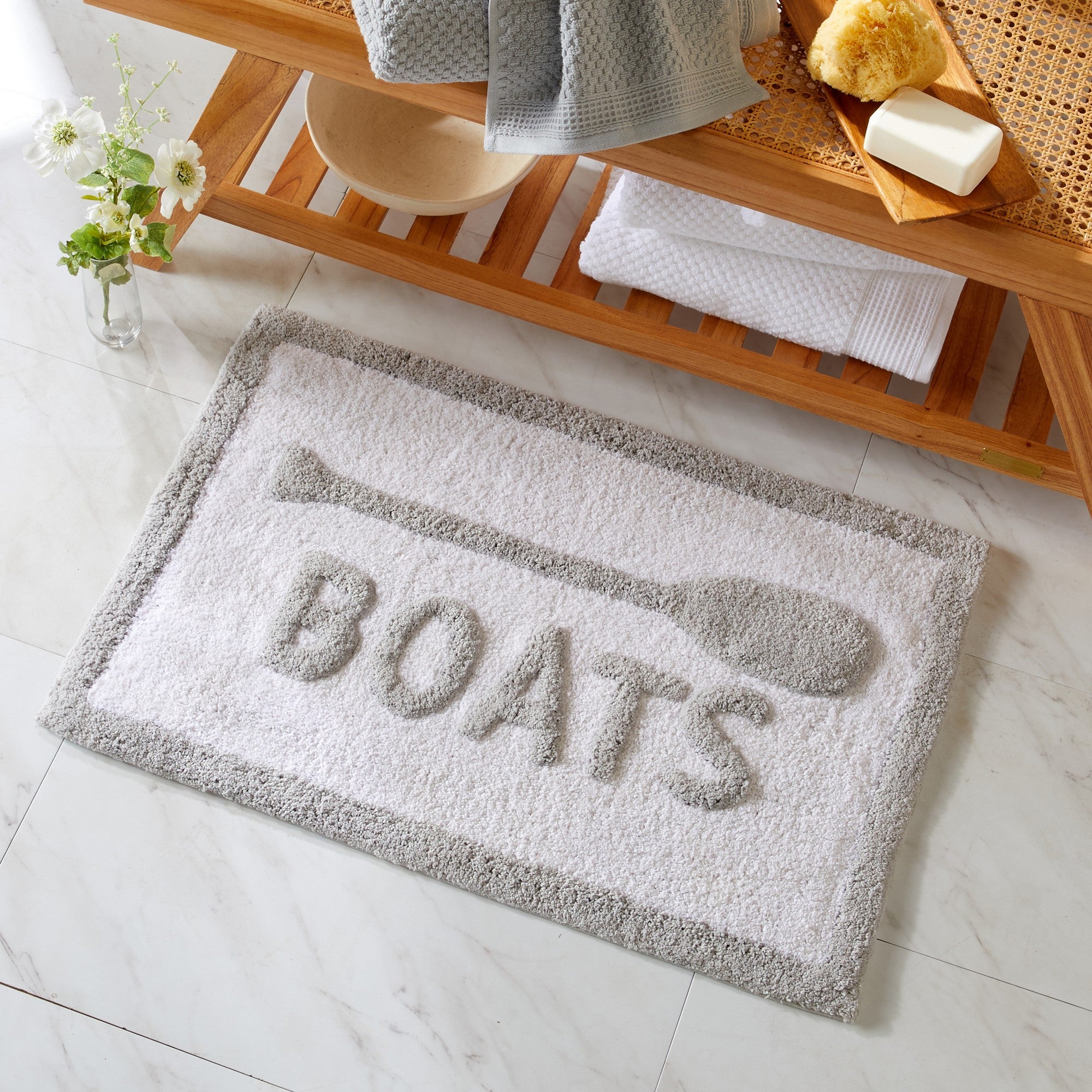 Gray doormat with 'BOATS' text on a bathroom floor, next to a wooden shelf with bath items.