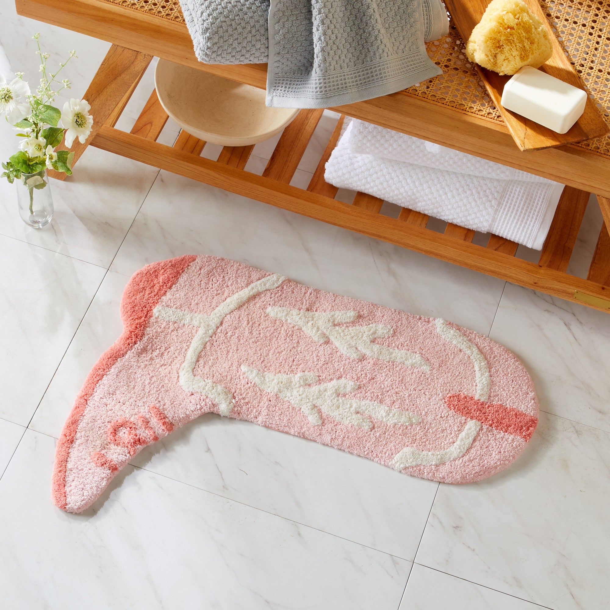 Flamingo-shaped bath mat on a tiled bathroom floor with a wooden shelf in the background.